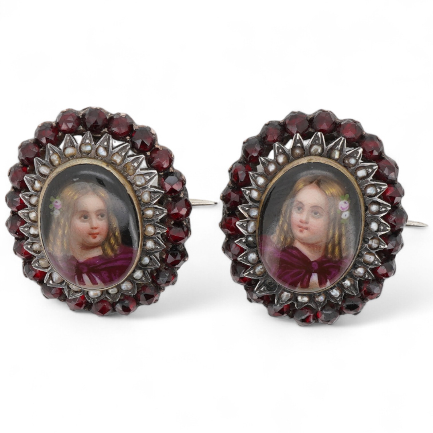 A pair of early 20th century yellow metal mounted brooches, with central oval panel painted with the bust of a lady and bordered by concentric rows of seed pearls and garnets (adapted), 24mm by 20mm.
